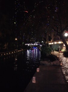 This is along San Antonio’s River Walk.  Candles in bags light our way, while lights strung overhead in the trees create an enchanted forest scene.  A brightly lit tour boat can be seen cruising down the river.